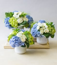 three white vases filled with blue and green flowers on a wooden tray next to each other