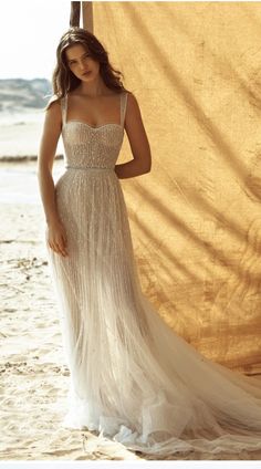 a woman in a white dress standing on the beach next to a tan curtain with her hands behind her head