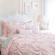 a bed with pink comforter and pillows in a white room next to a window