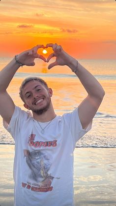 a man making a heart shape with his hands on the beach at sunset or sunrise