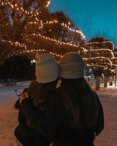 two people standing in the snow hugging each other with christmas lights on the trees behind them