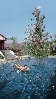 a woman floating in a swimming pool next to a christmas tree with decorations on it