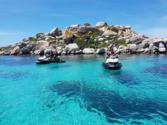 two people on jet skis in clear blue water near an island with large rocks