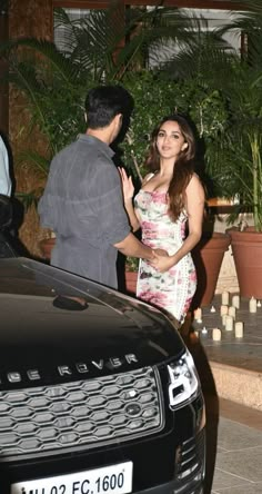 a man standing next to a woman in front of a black car and potted plants