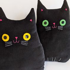 two black cat pillows with yellow eyes and green eyes on the front one has a pink nose