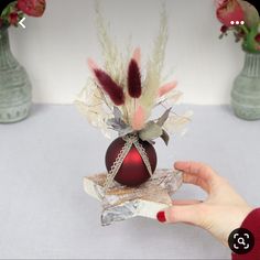 a hand is holding an ornament with feathers on it and flowers in the background