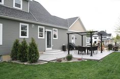 a patio with a table and chairs next to a gray house on the grass in front of it