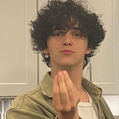 a man with curly hair and glasses standing in front of a white wall holding his hands together