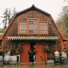 two people standing in the doorway of a wooden building with barrels on the outside and an awning over it