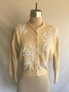 Lovely early 1960's hand beaded floral wool cropped cardigan! Wool is finely knit and beading features both white glass seed beads and faux pearl beads in a large flower pattern. Such a fine example of rockabilly and pin up vintage style! Cardigan is clean and is in overall great condition. There are a couple tiny mends at the bottom of the placket on the front near rib hem and at back right sleeve hear cuff. There is one tiny hole at center of left sleeve cuff and an even smaller hole at the ri Vintage Cream Sweater With Floral Embroidery, Vintage Cream Sweater For Spring, Vintage Cream Embroidered Cardigan, Vintage Embroidered Cream Cardigan, Fitted Cream Sweater With Floral Embroidery, Fitted Cream Sweater With Buttons, Vintage Beige Embroidered Sweater, Vintage Spring Sweater With Buttons, Vintage Embroidered Beige Sweater