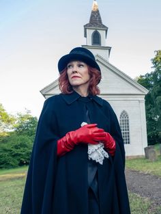 a woman in a black cape and red gloves is standing near a white church with a steeple