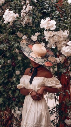 a woman wearing a white dress and hat standing in front of a bush with flowers