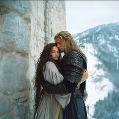 a man and woman hugging each other in front of a stone wall with mountains behind them