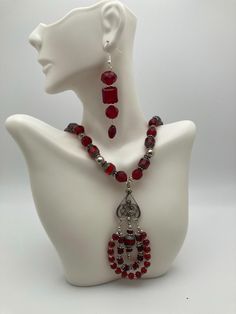 This striking 22" necklace and earrings set, primarily composed of red and silver beads. The necklace showcases faceted red beads with a deep, rich color, accented by silver spacers that add a touch of elegance. The centerpiece of the necklace is an ornate silver pendant with a teardrop shape, adorned with intricate designs and additional dangling red beads, giving it a dramatic flair. The matching earrings are equally bold, with a drop design featuring three faceted red beads in varying shapes and sizes, separated by silver accents. This set has a sophisticated and glamorous style, perfect for formal occasions or adding a pop of color to an outfit. Red Czech Glass Jewelry With Faceted Beads, Red Dangle Jewelry With Faceted Beads, Red Beaded Czech Glass Jewelry, Red Czech Glass Jewelry With Polished Beads, Red Ruby Beaded Necklaces With Faceted Details, Red Ruby Faceted Beaded Necklaces, Red Ruby Beaded Necklace With Faceted Details, Red Ruby Cut Beaded Necklaces, Red Beaded Ruby Jewelry