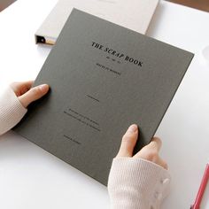 a person holding a book on top of a white table next to a red pen