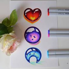 three different designs on paper next to some markers and a flower with pink flowers in the background