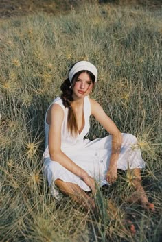 a young woman sitting in the grass wearing a white dress and headband with her hands on her knees