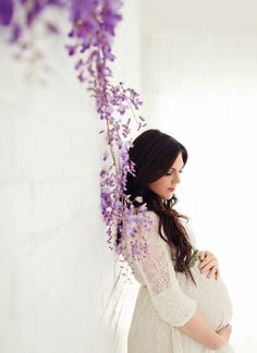 a pregnant woman standing next to a white wall with purple flowers on it's head