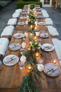 a long wooden table topped with white plates and greenery next to candles on top of snow covered ground