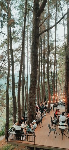 many people are sitting at tables in the woods by the water and some have lights on them