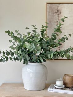 a white vase filled with green leaves sitting on top of a wooden table next to a candle