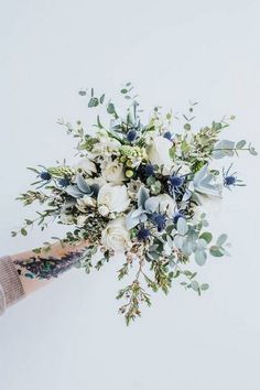 a person holding a bouquet of white and blue flowers with greenery on the stems