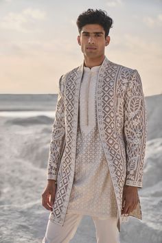 a man standing in front of the ocean wearing a white outfit and jacket with an intricate pattern on it