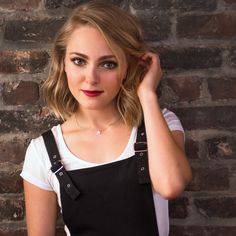 a woman with blonde hair and blue eyes posing in front of a brick wall holding her hand on her head