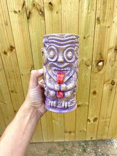 a hand holding up a cup with a face on the outside, and a wooden fence behind it