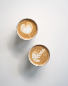 two cups filled with latte art on top of a white table next to each other