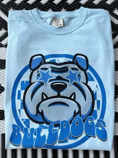 Get this popular new Bulldogs Mascot shirt and show off your school spirit in style!  Each shirt is printed in-house by me using high-quality materials on a Comfort Colors shirt, known for its exceptional comfort, style, and durability. Shown in Chambray. Even the glitter is printed! No need to worry about peeling glitter vinyl, or messy glitter flakes. Available in unisex sizes S to 3X, and youth sizes, we've got you covered regardless of your preferred fit. The Preppy Bulldogs Shirt features a Crew Neck Cotton Sublimation Design For School, Game Day Blue Pre-shrunk Shirt, Blue School Spirit Shirt With Graphic, Blue Pre-shrunk Shirt For Game Day, Blue Sublimation Print T-shirt For College, Blue School Spirit T-shirt With Sublimation Print, School Spirit Cotton Shirt With Graphic Print, Blue T-shirt With Sublimation Print For School Spirit, School Spirit Shirt With Graphic Print For Fans