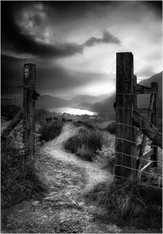 a black and white photo of a dirt path leading to a gate with mountains in the background