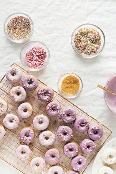 doughnuts and sprinkles sit on a cooling rack next to bowls of powdered sugar