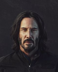 a man with long hair and beard wearing a black jacket looking at the camera while standing in front of a dark background