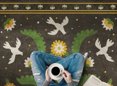 a woman sitting on the floor holding a coffee cup in front of her face with birds painted on the wall behind her