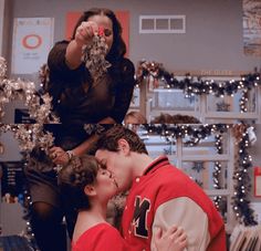 two young people are kissing in front of christmas decorations and lights, one is wearing a red jacket