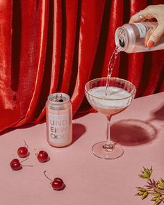 a person pouring liquid into a glass next to cherries on a pink table cloth