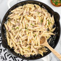 a skillet filled with pasta and meat on top of a marble countertop next to a wooden spoon