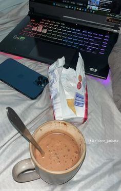 a cup of hot chocolate next to a laptop on a bed with a bag of chips