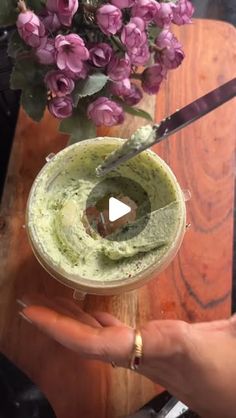 a person holding a cup filled with green liquid and purple flowers on top of a wooden table