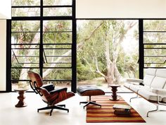 an eames lounge chair and ottoman in front of large open window with trees outside