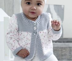 a baby wearing a knitted hat and cardigan is sitting on a white bench