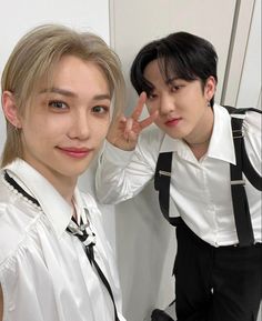 two young men standing next to each other in front of a white wall and wearing suspenders