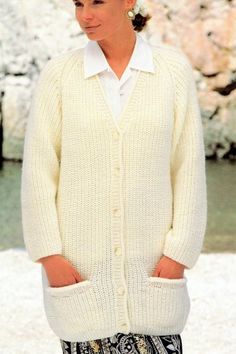 a woman wearing a white cardigan standing in front of some rocks and water with her hands in her pockets