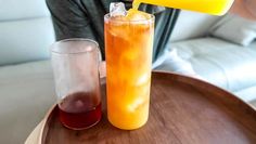 a person pouring orange juice into a tall glass on top of a wooden table next to a cup