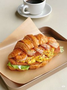 a croissant sandwich with shrimp and lettuce on it sits on a tray next to a cup of coffee