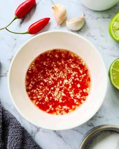 a white bowl filled with red sauce next to sliced limes and garlic on a marble counter