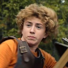 a young man with curly hair wearing an orange shirt and carrying a guitar case on his back