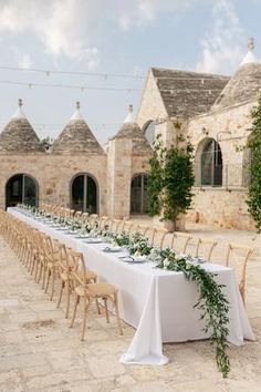 a long table is set up outside with white linens and greenery on it