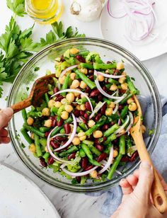 a person holding a wooden spoon in a bowl filled with beans, onions and broccoli